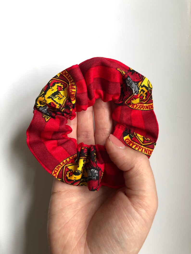 May include: Red and yellow fabric hair scrunchie with a repeated pattern of the Gryffindor house crest from the Harry Potter series. The crest features a lion, a house name, and a diagonal stripe.
