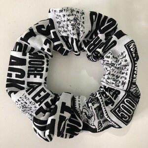 May include: Black and white scrunchie with a newspaper print design. The scrunchie features text such as "MORE PLEASE" and "500".