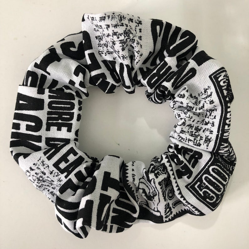May include: Black and white scrunchie with a newspaper print design. The scrunchie features text such as "MORE PLEASE" and "500".
