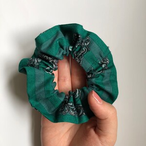 May include: Green fabric hair scrunchie with a white and gray pattern. The pattern features a crest with a snake and a crown.