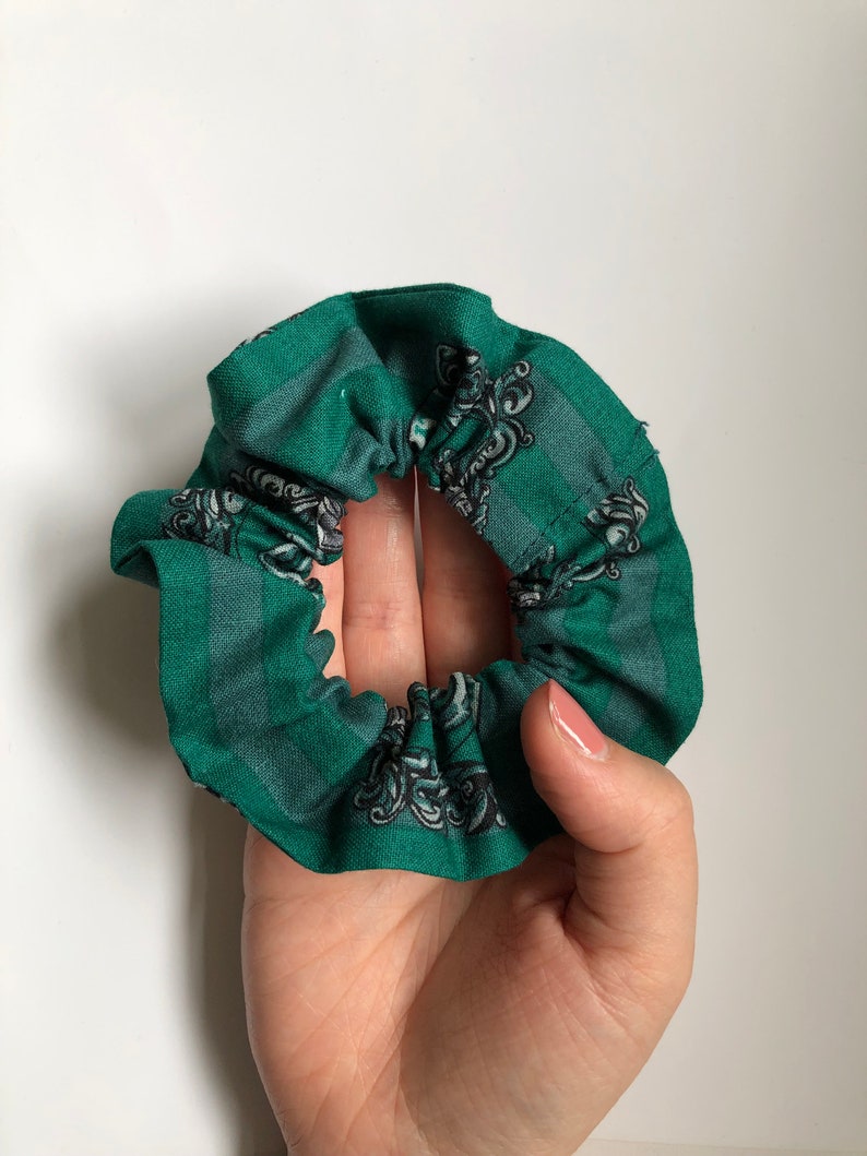 May include: Green fabric hair scrunchie with a white and gray pattern. The pattern features a crest with a snake and a crown.