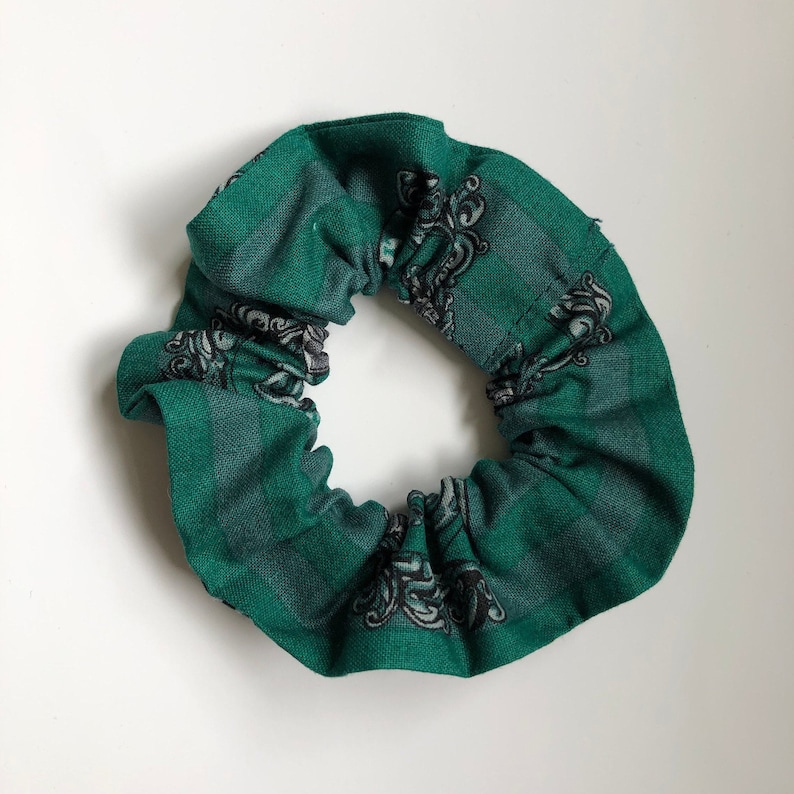 May include: Green fabric hair scrunchie with a black and white pattern. The pattern features a repeating design of a crest with a stylized animal.