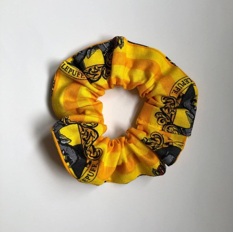 May include: Yellow and black hair scrunchie with a checkered pattern and the Hufflepuff crest. The crest features a badger, a  "H" and the words "Hufflepuff" on a yellow background.