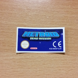 Puede incluir: Una etiqueta de cartucho de juego Game Boy Advance azul y blanca con el texto "METROID ZERO MISSION" y el logotipo de Nintendo. La etiqueta también incluye una marca CE y el código del juego "AGB-BMXP-EUR".