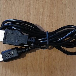 Può includere: Cavo di ricarica USB nero con connettore USB-A e connettore micro-USB. Il cavo è arrotolato e fissato con una fascetta nera. I connettori sono neri e argentati.