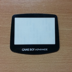 Puede incluir: Una pieza de repuesto de pantalla negra para Game Boy Advance. La pantalla es transparente y tiene un marco negro con las palabras "GAME BOY ADVANCE" impresas en blanco.