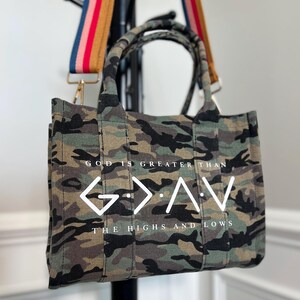 May include: A camouflage print tote bag with two top handles and a detachable, multi-colored striped shoulder strap. The front features the white text "GOD IS GREATER THAN" above a "G > ^ V" symbol and "THE HIGHS AND LOWS" below.