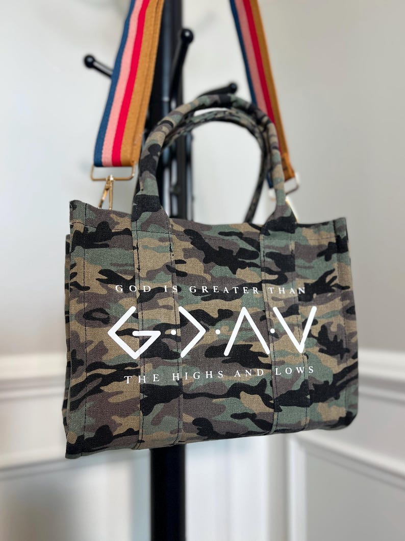 May include: A camouflage print tote bag with two top handles and a detachable, multi-colored striped shoulder strap. The front features the white text "GOD IS GREATER THAN" above a "G > ^ V" symbol and "THE HIGHS AND LOWS" below.