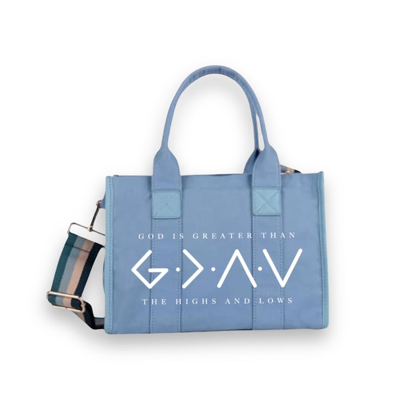 May include: A light blue canvas tote bag with two top handles and a detachable striped shoulder strap. The front features the white text "GOD IS GREATER THAN THE HIGHS AND LOWS" along with the corresponding G>&and;<V symbol printed in white.
