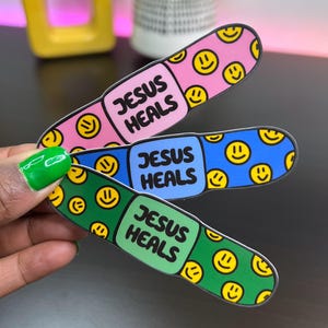May include: Three colorful band-aid stickers with the text "Jesus Heals" and smiley faces. The band-aids are pink, blue, and green.