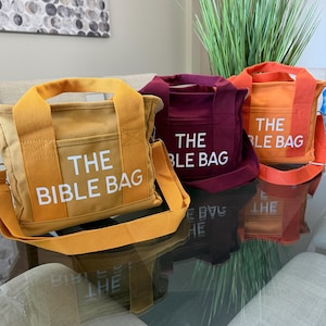 May include: Three canvas tote bags in shades of yellow, burgundy, and orange, each with a matching shoulder strap and the text "THE BIBLE BAG" printed in white. The bags are displayed on a reflective glass surface.