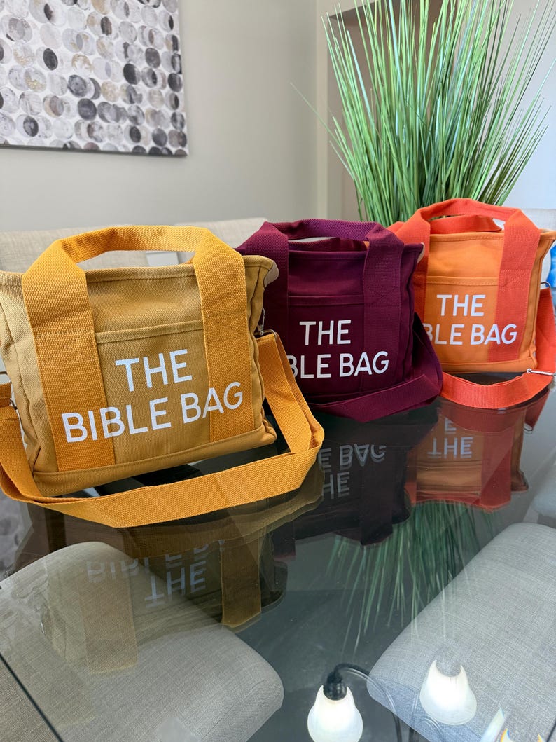 May include: Three canvas tote bags in shades of yellow, burgundy, and orange, each with a matching shoulder strap and the text "THE BIBLE BAG" printed in white. The bags are displayed on a reflective glass surface.