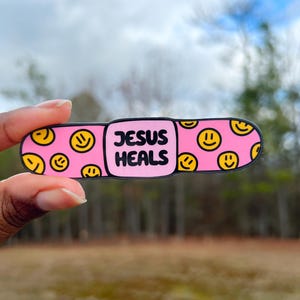 May include: Pink band-aid with a smiley face pattern and the text "JESUS HEALS" in black.