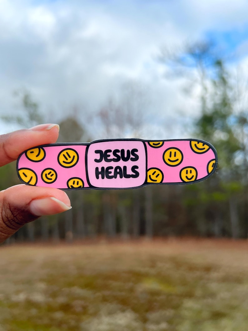 May include: Pink band-aid with a smiley face pattern and the text "JESUS HEALS" in black.