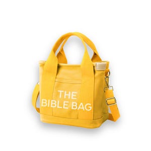 May include: A yellow canvas tote bag with the text "THE BIBLE BAG" printed in white. The bag has a shoulder strap and two handles. It features a small front pocket and a beige leather patch near the top.