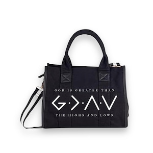 May include: A black canvas tote bag with two top handles and a detachable shoulder strap. The front features white text that reads GOD IS GREATER THAN and THE HIGHS AND LOWS, with the G > ^ < V symbol printed in the center between the lines.