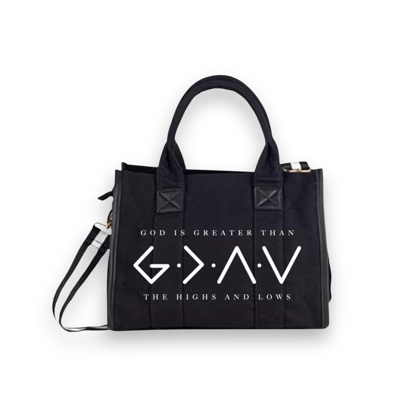 May include: A black canvas tote bag with two top handles and a detachable shoulder strap. The front features white text that reads GOD IS GREATER THAN and THE HIGHS AND LOWS, with the G > ^ < V symbol printed in the center between the lines.