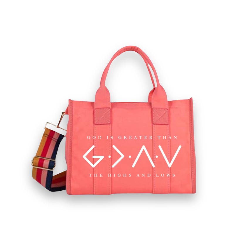 May include: A coral-colored tote bag featuring the white text GOD IS GREATER THAN THE HIGHS AND LOWS and a G>^<V symbol. The bag includes two top handles and a detachable shoulder strap with a multi-colored striped pattern in red, yellow, and blue.