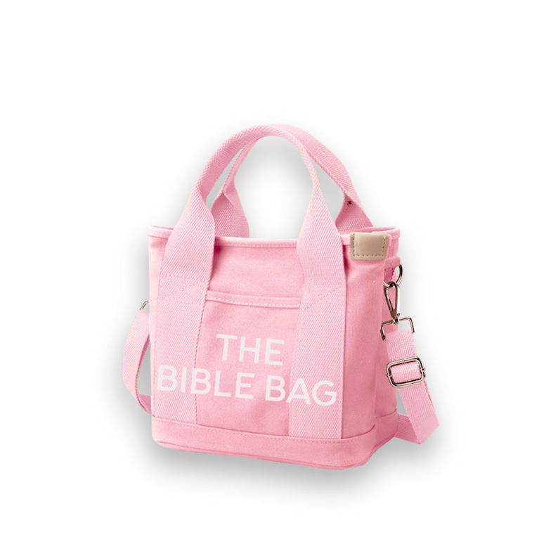 May include: A pink canvas tote bag with the text "THE BIBLE BAG" in white. The bag has two handles and a detachable shoulder strap. It features a small front pocket and a light brown tag.