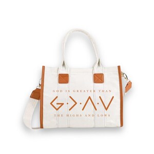 May include: A white canvas tote bag with brown leather-like trim and handles. The front features the brown text "GOD IS GREATER THAN THE HIGHS AND LOWS" and the symbol "G > ^ < V". It includes a detachable shoulder strap with matching brown accents.