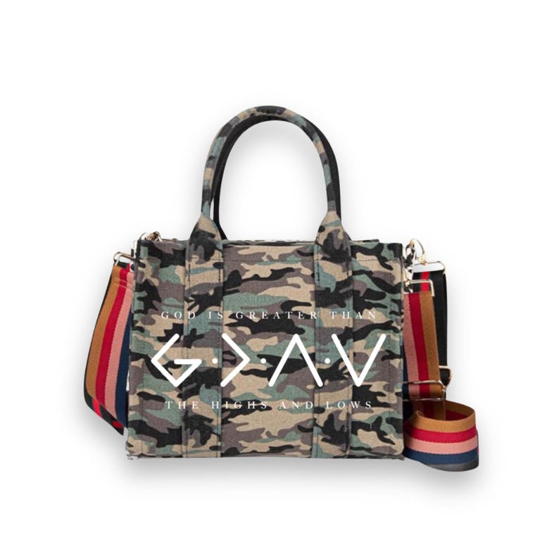 May include: A camouflage print tote bag with two top handles and a detachable, multi-colored striped shoulder strap. The front features the white text "GOD IS GREATER THAN THE HIGHS AND LOWS" with the symbol "G > ^ v" printed in the center.