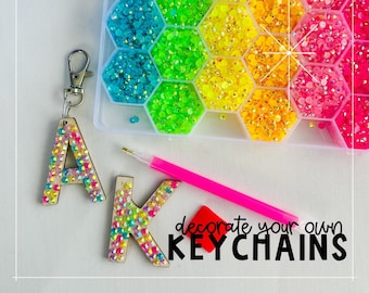 May include: A craft kit for decorating keychains. The kit includes a container of colorful rhinestones, wooden letters, a pink stylus, and the text "decorate your own keychains". The letters "A" and "K" are shown decorated with rhinestones.