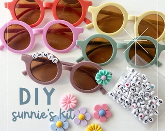 May include: DIY sunnies kit with colorful round sunglasses, flower charms, and letter beads. The sunglasses are in various colors, including pink, green, brown, and yellow. The flower charms are in shades of pink, blue, and yellow. The letter beads are white with black lettering.