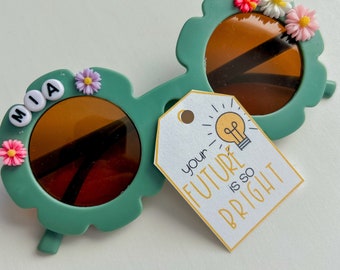 May include: Green plastic sunglasses with a flower design and the name "MIA" spelled out in white letters. A white tag with a yellow lightbulb and the text "your future is so bright" is attached to the sunglasses.