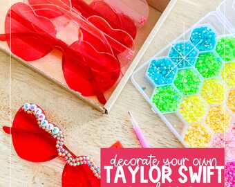 May include: Red heart-shaped sunglasses with the words "SWIFTIE" spelled out in colorful beads. A container of colorful rhinestones and a pink tool are also pictured. The text "decorate your own TAYLOR SWIFT sunglasses" is written in white letters on a pink background.