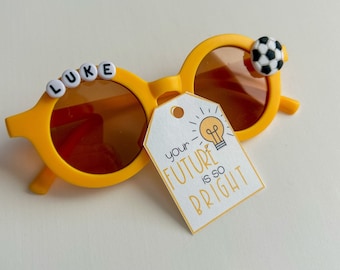 May include: Yellow sunglasses with a soccer ball charm and the name "LUKE" spelled out in white beads. A white tag with the text "your future is so bright" and a lightbulb graphic is attached to the sunglasses.