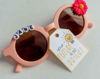 May include: Peach-colored sunglasses with brown lenses, featuring the name "JANE" spelled out in white letter beads. A pink flower adorns the frame, and a tag reads "your FUTURE is so BRIGHT."
