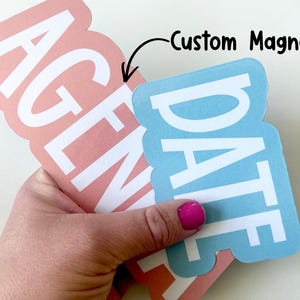 May include: Custom magnets in the image. One is peach-colored with the word "AGENDA" in white, and the other is light blue with "DATE" in white. The text "Custom Magnets" is written in black.
