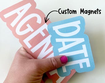 May include: Custom magnets in the image. One is peach-colored with the word "AGENDA" in white, and the other is light blue with "DATE" in white. The text "Custom Magnets" is written in black.