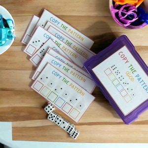 May include: A set of six printable game cards with the text "Copy the Pattern" and a pattern of dots on each card. The cards are stacked on top of each other. A purple plastic container with the text "Copy the Pattern dice" and a pattern of dots on a die is in the background. There are four white dice on a wooden surface.