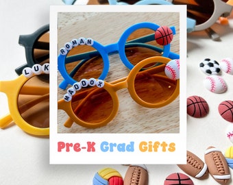 May include: Pre-K grad gifts featuring personalized blue and yellow sunglasses with names "Roman", "Maddox", and "Luk" decorated with sports-themed charms. The charms include a basketball, baseball, football, soccer ball, and volleyball.