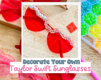 May include: Red heart-shaped sunglasses decorated with colorful rhinestones and the word "SWIFTIE" in white letters. The sunglasses are on a wooden surface with a container of colorful rhinestones in the background. The text "Decorate Your Own Taylor Swift Sunglasses" is at the bottom of the image.