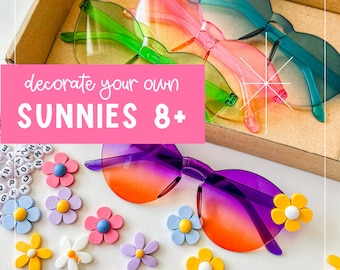 May include: A collection of colorful sunglasses with various frame colors, including green, pink, and blue, and gradient lenses. The image also features decorative flower embellishments and the text "decorate your own SUNNIES 8+".