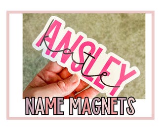 May include: A white magnet with the name "Ansley" written in pink letters. The magnet is being held by a hand. The text "NAME MAGNETS" is written in pink letters at the bottom of the image.