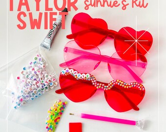 May include: A DIY craft kit to personalize heart-shaped sunglasses with colorful beads and letters. The kit includes red and pink sunglasses, a bag of letter beads, a bag of colorful beads, a tube of glue, a pink pen, and a red square.