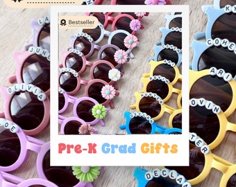 May include: A collection of colorful, round sunglasses with personalized names, such as Olivia, June, and Hunter, and a flower embellishment on each pair. The sunglasses are arranged in a row on a wooden surface. The text "Pre-K Grad Gifts" is displayed in a colorful banner.