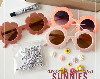 May include: A craft kit with flower-shaped sunglasses in shades of pink and peach. The kit includes letter beads, decorative flowers, and glue. The text "decorate your own SUNNIES" is also visible.