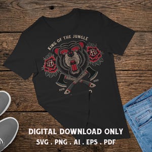 May include: A black t-shirt featuring a graphic of a snarling bear head centered above two crossed axes. The design includes red roses on either side of the bear, with the text "KING OF THE JUNGLE" arched above and "ESTD 2021" below.
