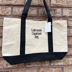 May include: A beige canvas tote bag with black handles and a black bottom. The bag has the text "Ludicrously Capacious Bag" embroidered on the front.
