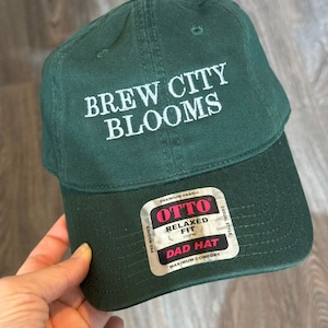 May include: A green dad hat with the text "Brew City Blooms" embroidered on the front. The hat has a relaxed fit and is made of premium fabric. The label on the side reads "Otto Relaxed Fit OSFM Dad Hat Maximum Comfort Casual Style Pre-Washed Premium Fabric"