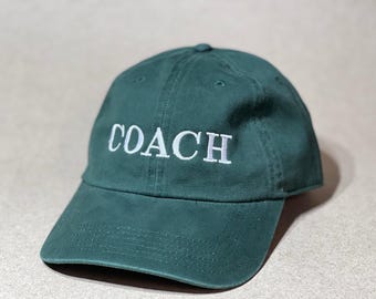 以下が含まれることがあります： 前面に白で「COACH」の文字が刺繍された濃い緑色の野球帽。つばがカーブしており、丸みを帯びたクラウンで、カジュアルな服装に適しています。