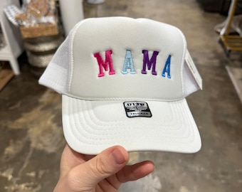 May include: White trucker hat with a mesh back. The front of the hat has the word "MAMA" embroidered in pink, blue, and purple letters.