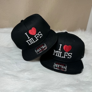 May include: Two black baseball caps with white embroidered text and a red heart. The text on the caps reads "I love DILFS" and "I love MILFS".