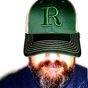 May include: A green and white trucker hat with a large, embroidered green "R" on the front. The hat has a dark green bill and a mesh back. The hat is worn by a person with a beard.