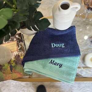 May include: Two embroidered towels, one navy blue with "Doug" in light green, and one light green with "Mary" in dark green. The towels are folded and stacked on a glass table, with a white ceramic pitcher and a potted plant in the background.
