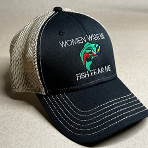 Op de afbeelding: Zwarte en beige truckercap met een wit geborduurde vis en de tekst "Women Want Me Fish Fear Me"